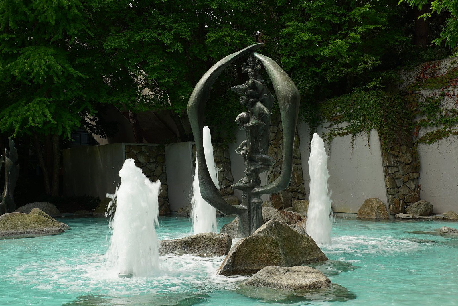 The fountain in its wading pool era, turquoise water with boulders and water jets, lush green trees