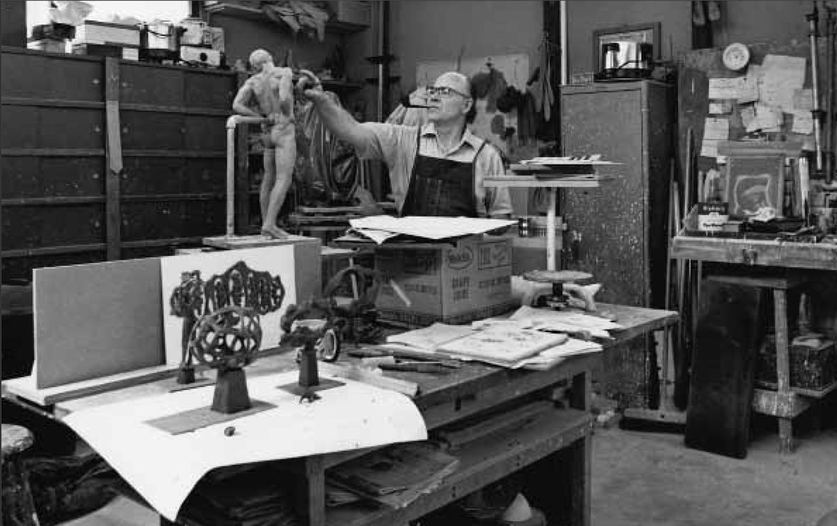 DuPen in his studio surrounded by bronzes and tools, holding a figure at arm's length